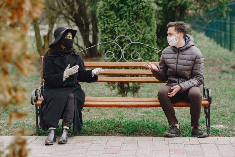 Two people wearing masks sit and converse on opposite ends of a park bench.